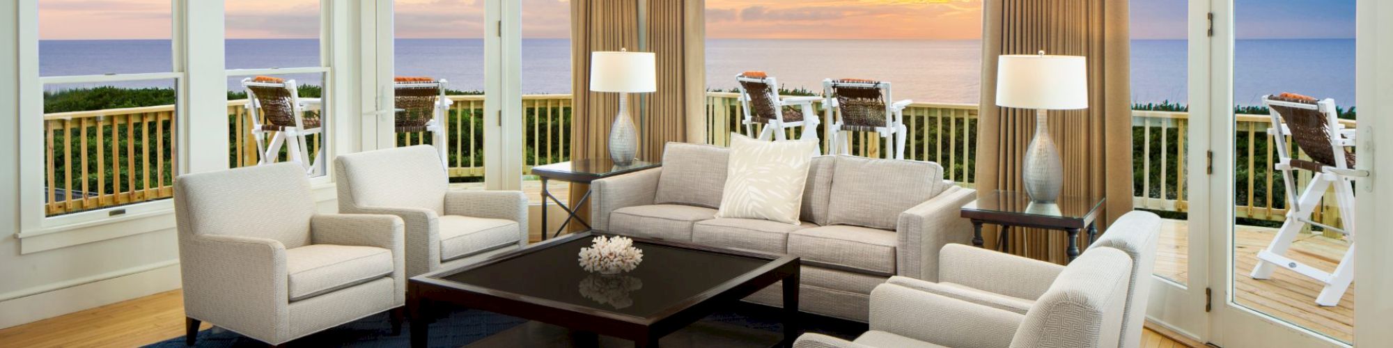 A cozy living room with beige sofas, a dark coffee table on a blue rug, and large windows offering an ocean view at sunset, complete with a balcony.