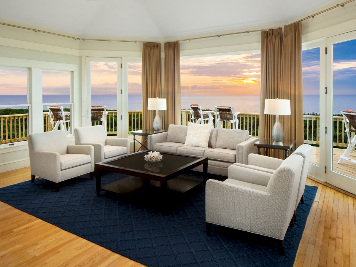 A cozy living room with white furniture, a dark coffee table on a navy rug, and large windows revealing an ocean view at sunset.