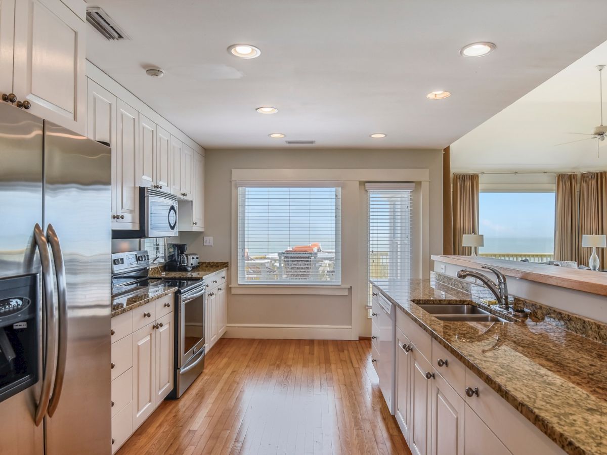 A modern kitchen with stainless steel appliances, granite countertops, and wooden flooring, overlooking a living area with large windows and ocean views.