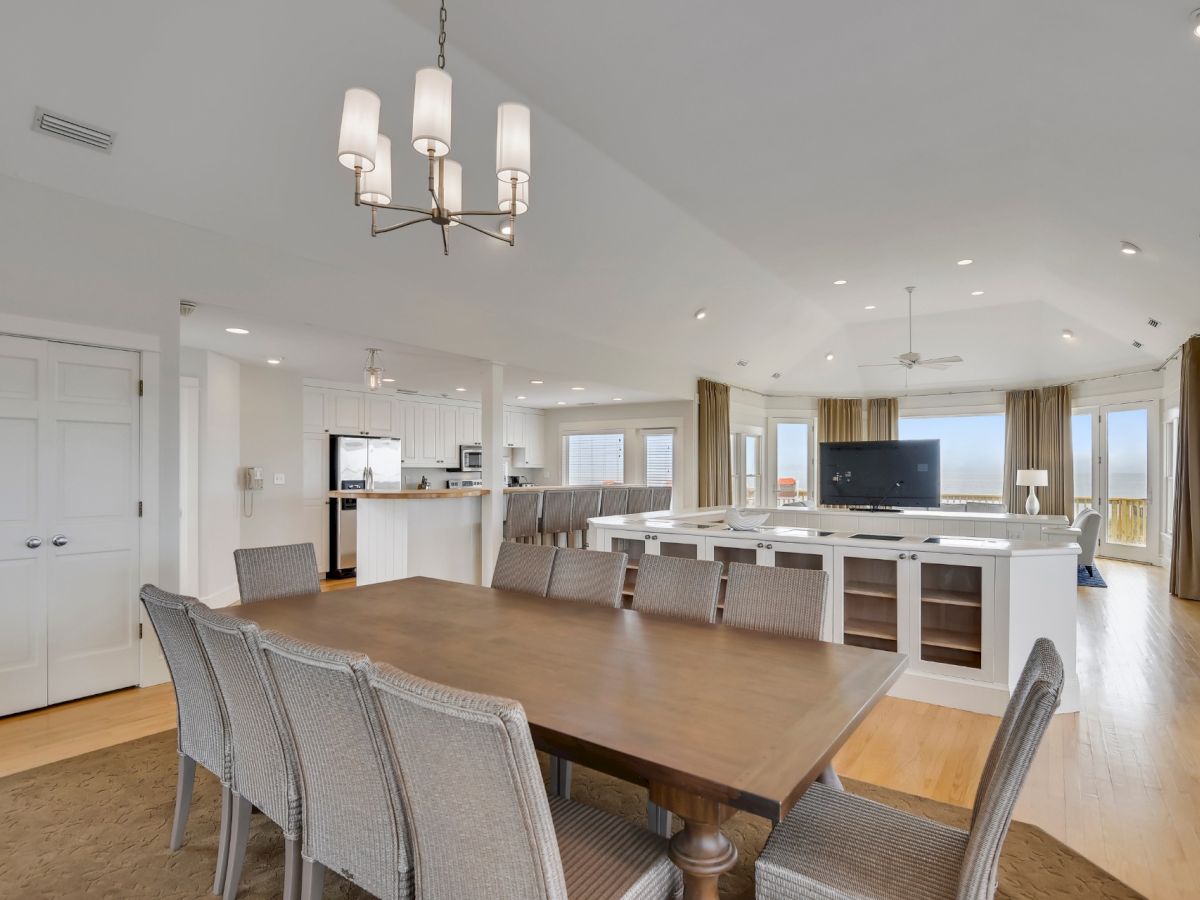 A spacious open-plan living area with a dining table, six chairs, a living room with a TV, and a kitchen with white cabinets and island seating.