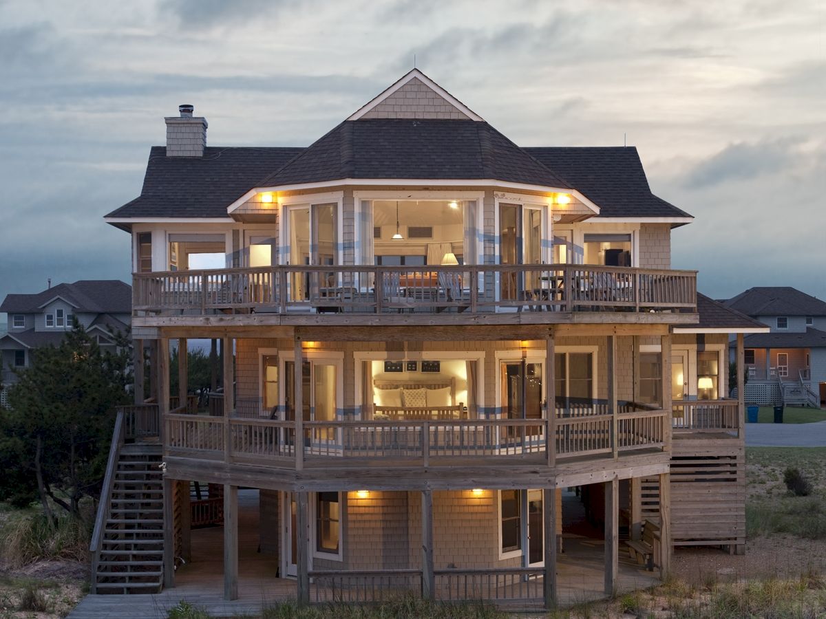 A three-story house with multiple balconies is lit up as dusk approaches, situated in a grassy area with a cloudy sky overhead.