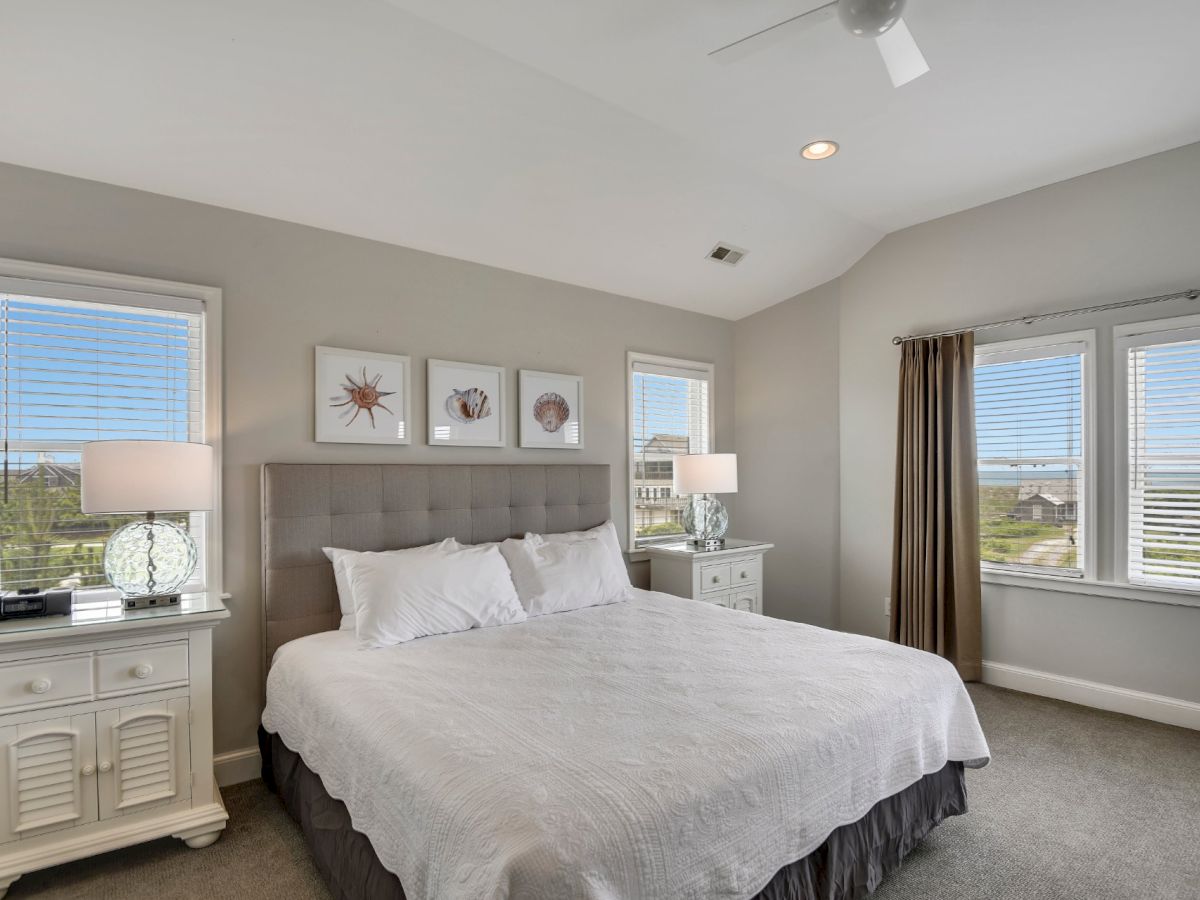A modern bedroom with a large bed, white linens, two nightstands, lamps, three wall art pieces, a ceiling fan, beige walls, and windows with a scenic view.