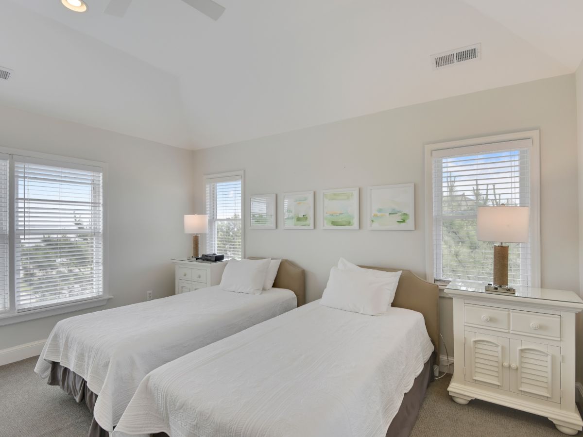 A bedroom with two twin beds, white bedding, two bedside tables with lamps, wall art above the beds, and windows with blinds letting in natural light.