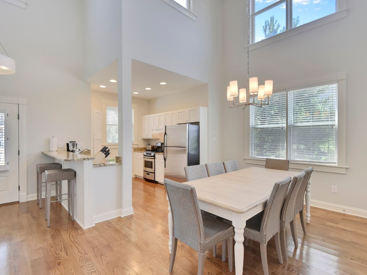 Modern kitchen and dining area with wooden floors, white furniture, stainless steel appliances, and large windows letting in natural light.
