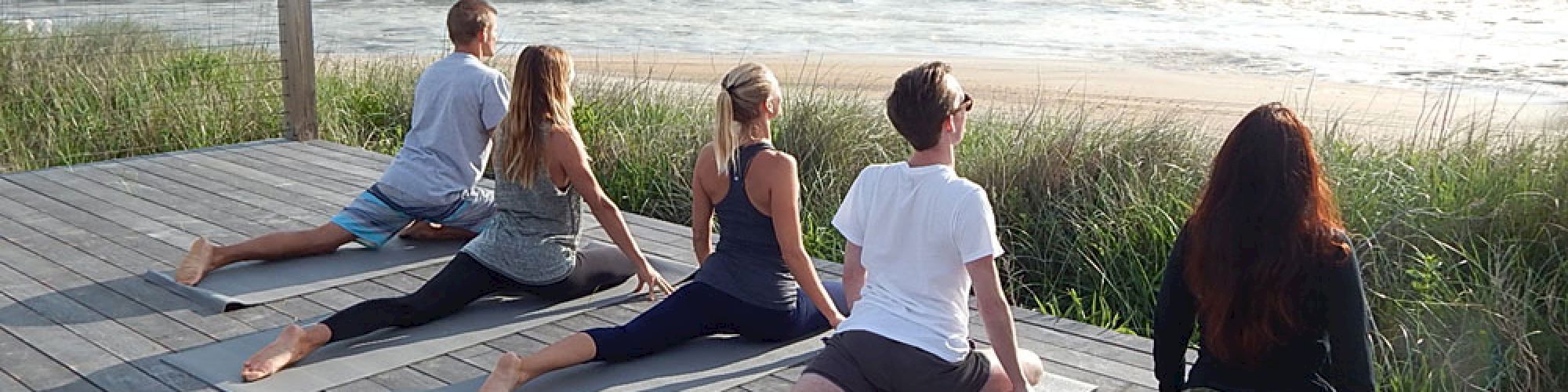 A group of five people practicing yoga on mats by the ocean, enjoying a scenic, serene view of the sea and sky, on a wooden deck.