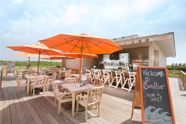 An outdoor beachside bar and dining area with orange umbrellas, wooden tables and chairs, and a welcome sign saying 