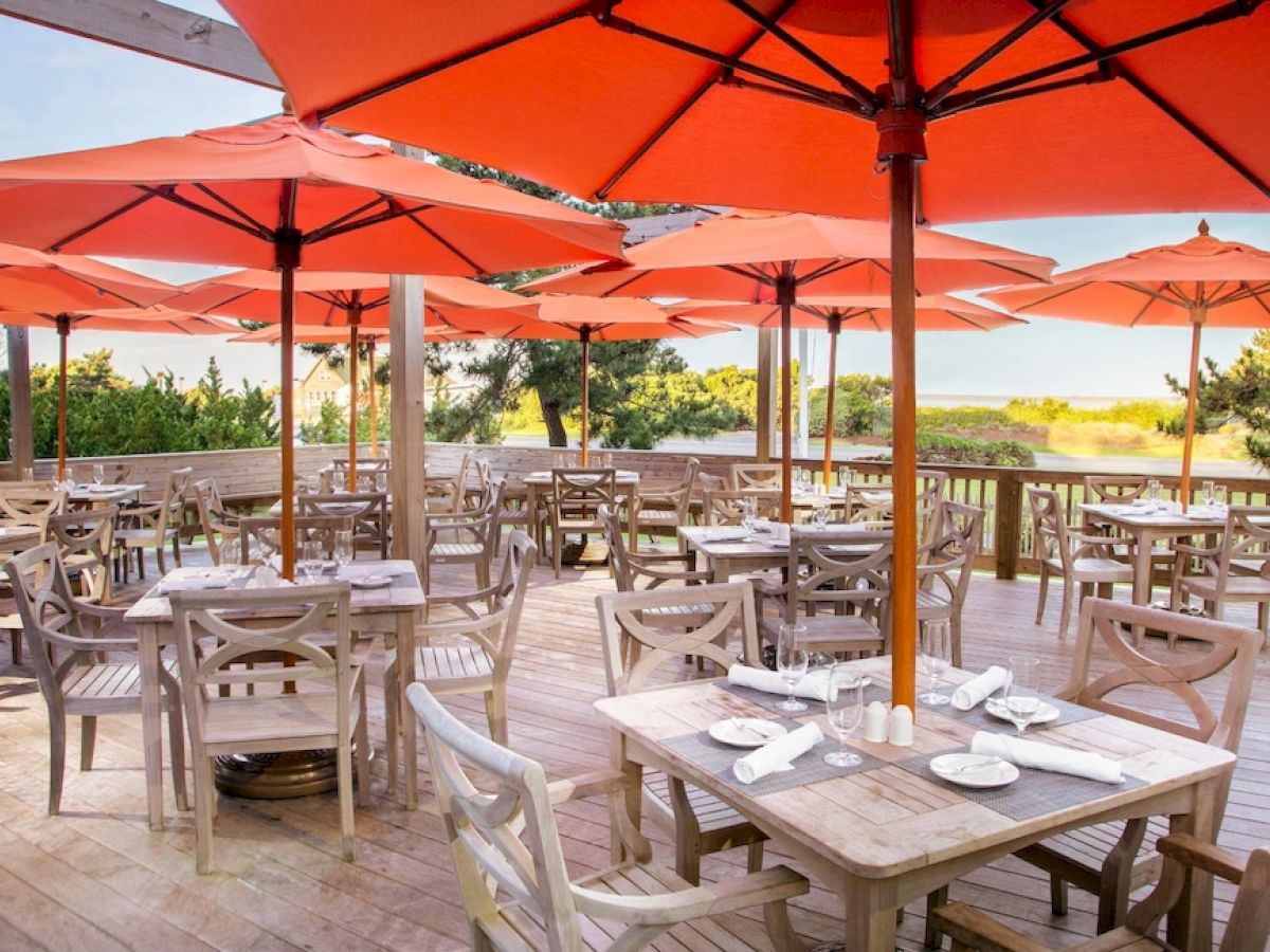 An outdoor restaurant with wooden tables and chairs, each shaded by large orange umbrellas. The setting overlooks a scenic, green landscape.