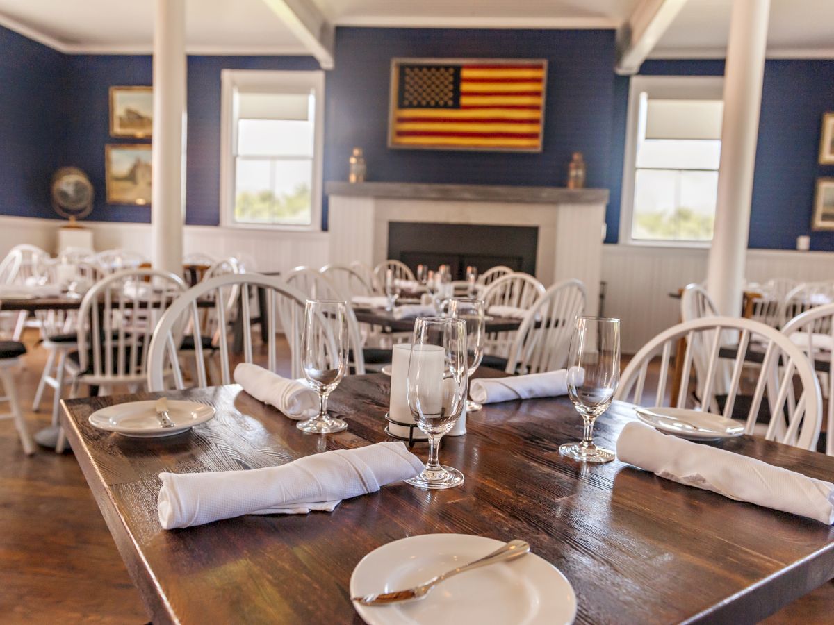 The image shows a neatly set dining area with wooden tables, white chairs, table settings, and an American flag on the wall behind.