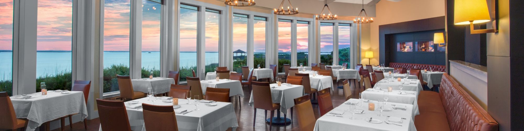 A spacious, elegant restaurant with large windows offering a sunset view, well-set tables with white cloths, brown chairs, and cozy lighting.
