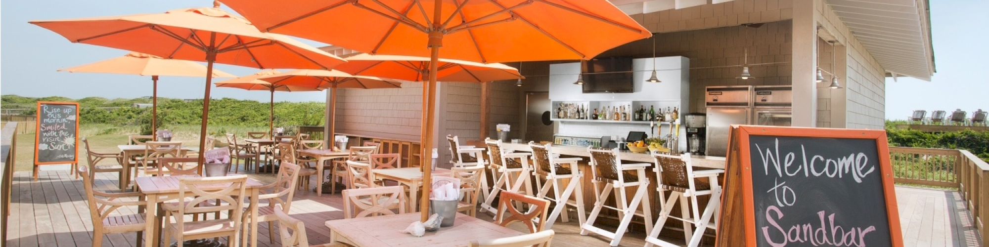 An outdoor café with orange umbrellas, wooden tables and chairs, and a chalkboard sign stating 