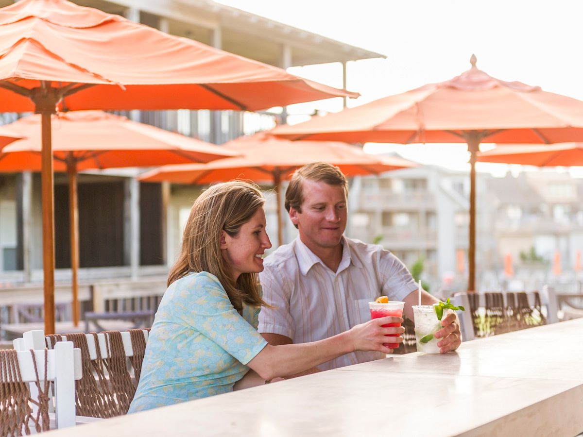 A man and a woman are sitting at an outdoor bar with orange umbrellas, enjoying colorful drinks together, and surrounded by a bright atmosphere.