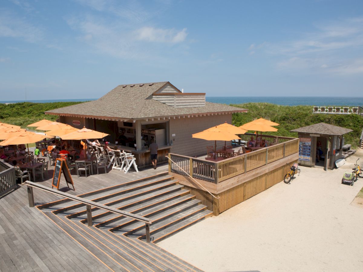 An outdoor beachside café with orange umbrellas on a deck, surrounded by wooden walkways with the ocean visible in the background, under a clear sky.