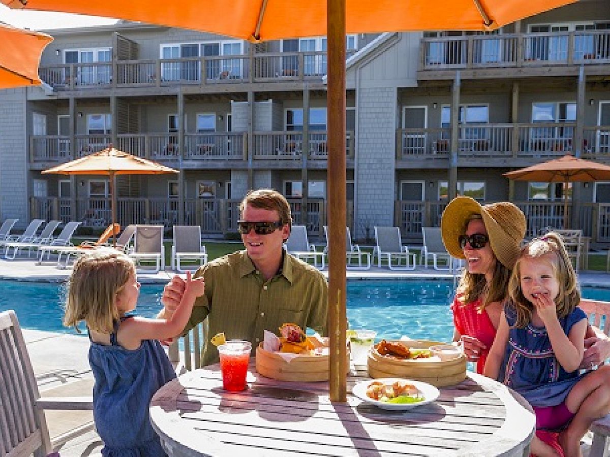 A family of four is dining at an outdoor table near a pool, with food and drinks on the table, under orange umbrellas, and resort buildings in the background.