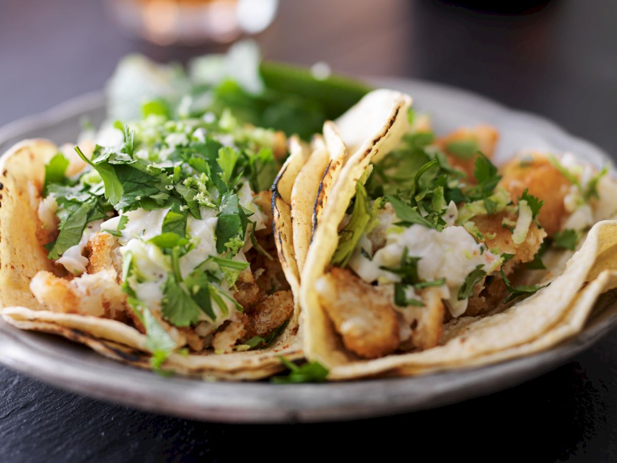 The image shows two tacos filled with what appears to be fried fish, topped with chopped cilantro and a white sauce, served on a plate.