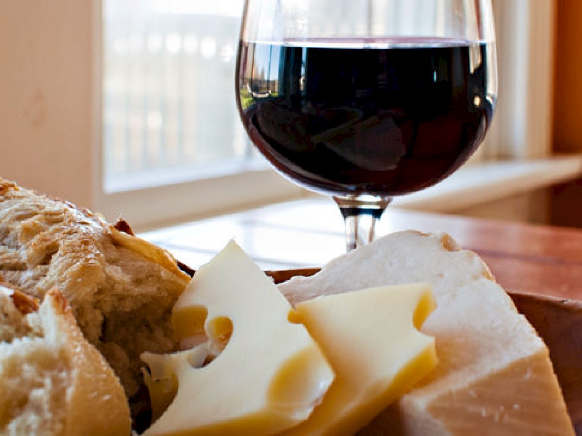 The image shows a glass of red wine next to a wooden plate with bread and slices of cheese. The background features a bright, sunlit window.