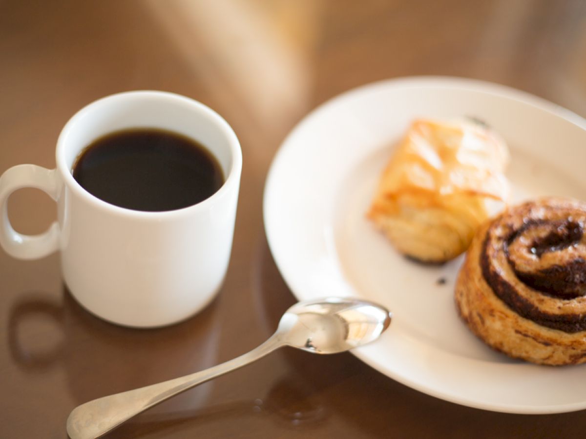 A cup of black coffee sits beside a white plate with a croissant and a cinnamon roll, along with a silver spoon.