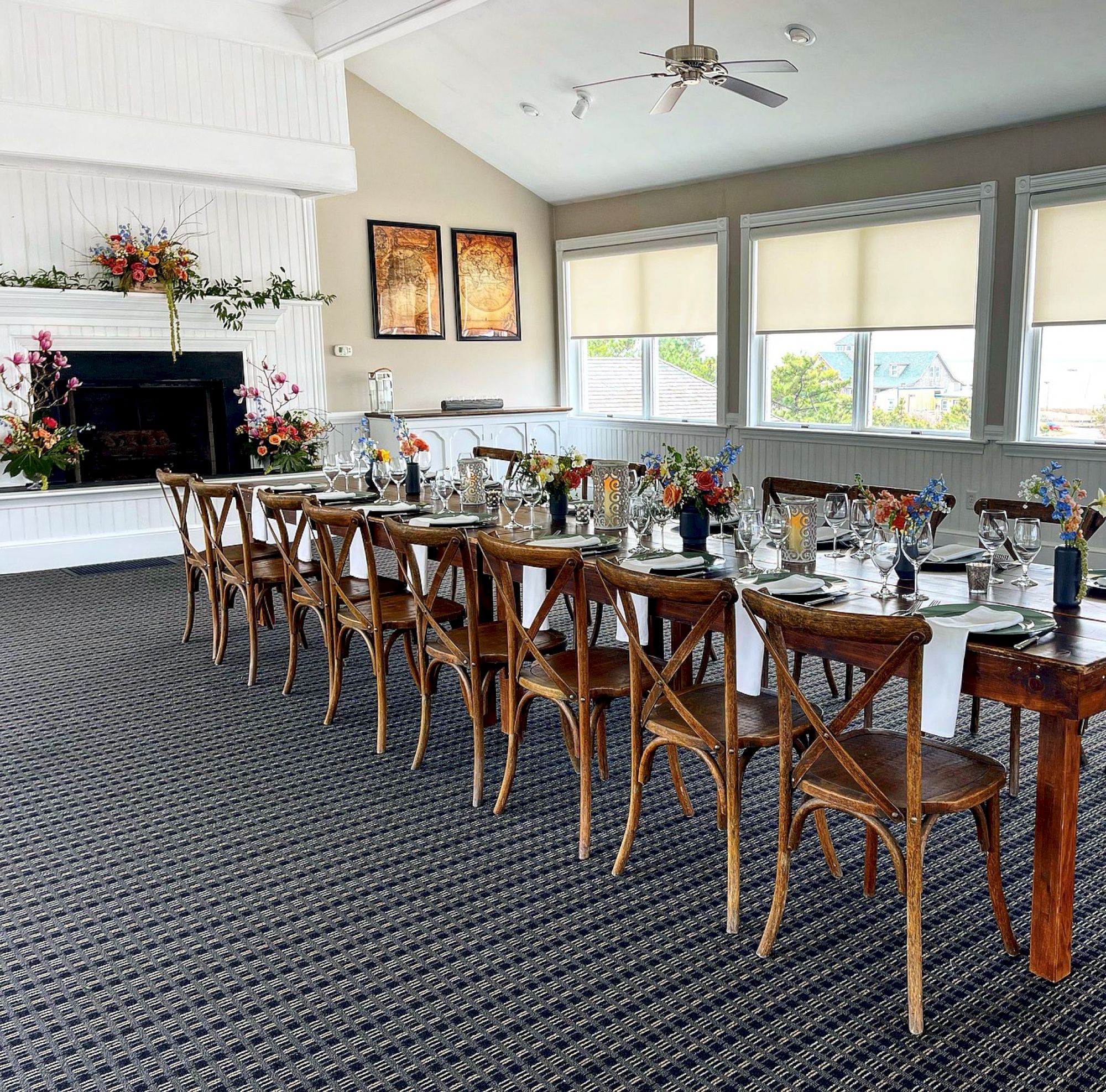 A long dining table with wooden chairs, set with glassware and floral arrangements, is in a bright, carpeted room with large windows and framed art.
