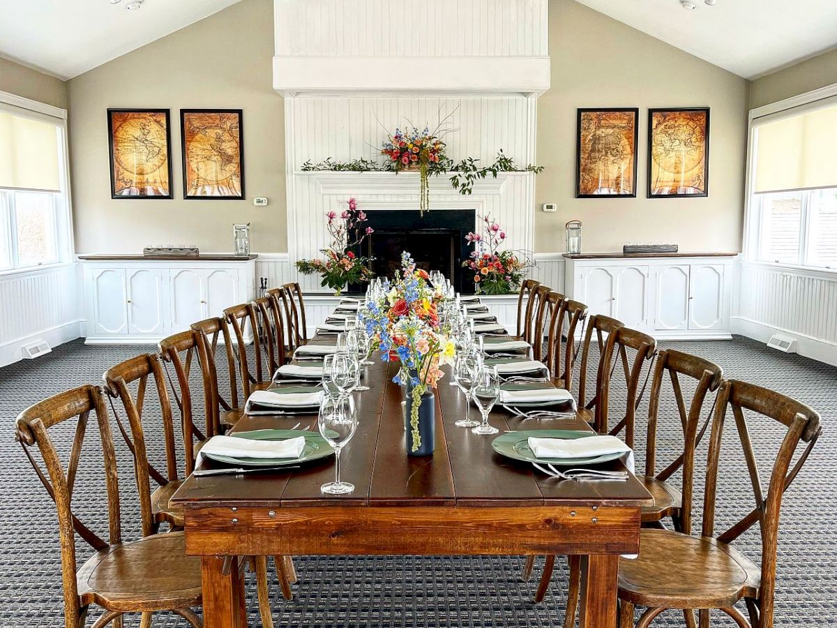 A long, elegant dining table is set in a well-lit room with wooden chairs, place settings, and a floral centerpiece, surrounded by paintings and decor.