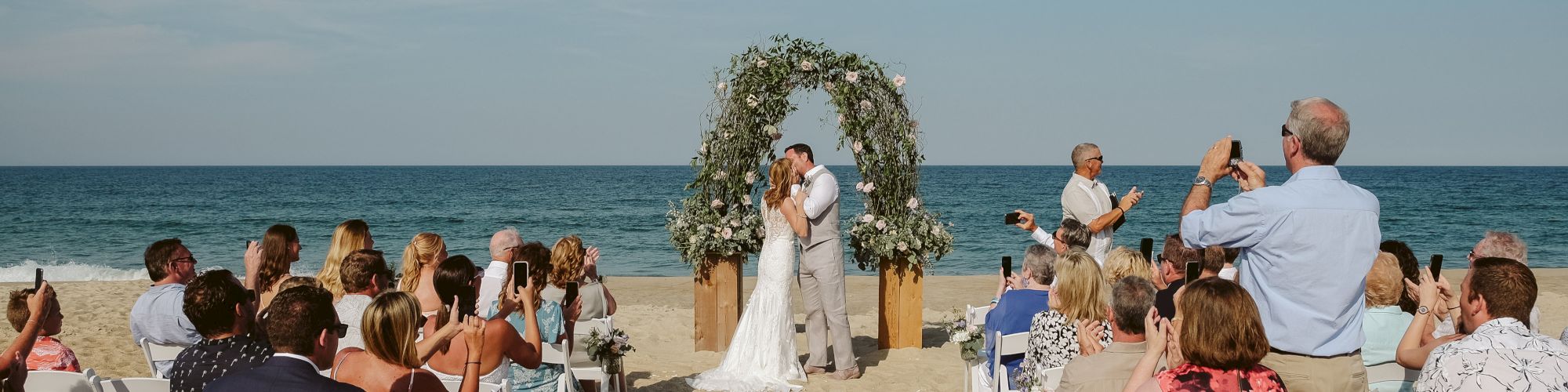 A couple is having a beach wedding ceremony, standing under a floral arch, surrounded by guests seated on white chairs, with the ocean in the background.