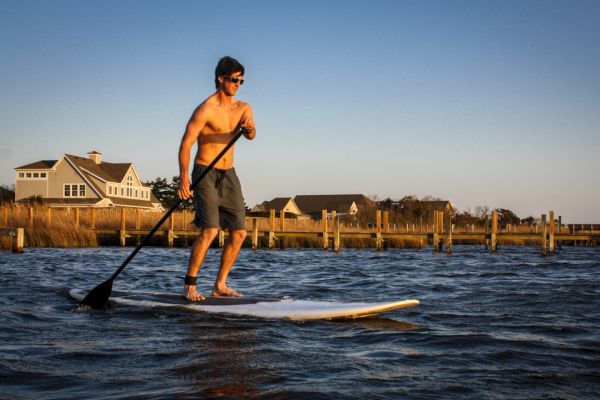 A person is paddleboarding on a body of water near some houses and a dock under a clear sky.