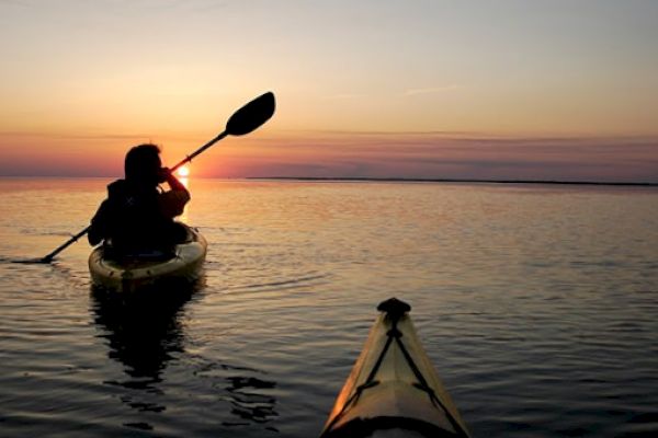 A person is kayaking on a calm body of water during sunset, with another kayak visible in the foreground, creating a peaceful and picturesque scene.