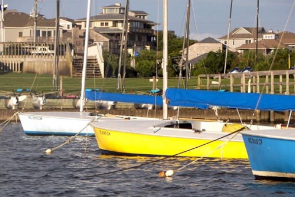 The image shows a row of sailboats docked in a marina, with houses and greenery visible in the background, providing a peaceful waterfront scene.