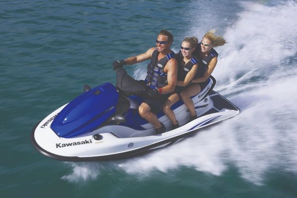 Three people riding a blue and white Kawasaki jet ski on water, all wearing life jackets and sunglasses, with water spray visible.