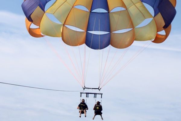 Two people are parasailing, harnessed to a colorful parachute against a backdrop of blue sky and clouds.