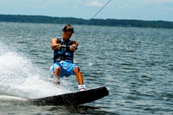 The image shows a person wakeboarding on a lake, wearing a life vest and blue shorts, being pulled by a rope while riding over the water.