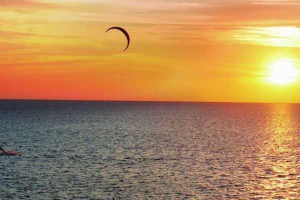 A person is kiteboarding on the ocean with a vibrant sunset in the background, casting an orange-yellow glow over the water.