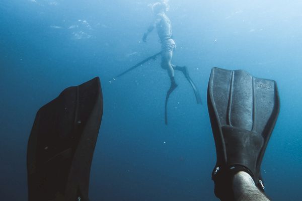 A view from underwater shows someone wearing flippers, with another diver swimming above them in the blue ocean.