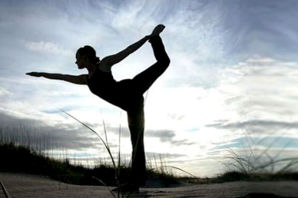 A person in silhouette is performing a yoga pose outdoors against a backdrop of a partly cloudy sky and grassy terrain.