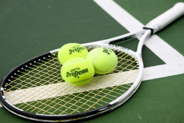 A tennis racket lies on a tennis court with three yellow tennis balls on top of it, positioned near the white boundary line.