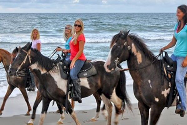 Four people are riding horses along a sandy beach with ocean waves in the background, enjoying a scenic ride.