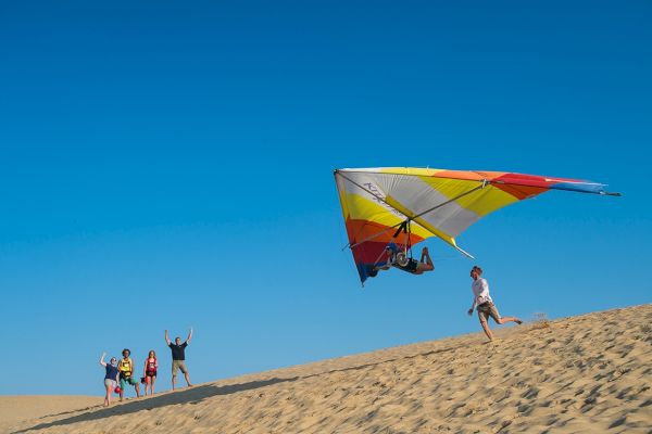 People on a sandy hill, with one person hang gliding while others cheer and one runs alongside, under a clear blue sky.