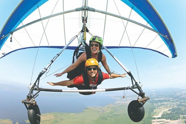 Two people are tandem hang gliding over a scenic landscape with helmets on, smiling and enjoying the experience of flying in the open sky.