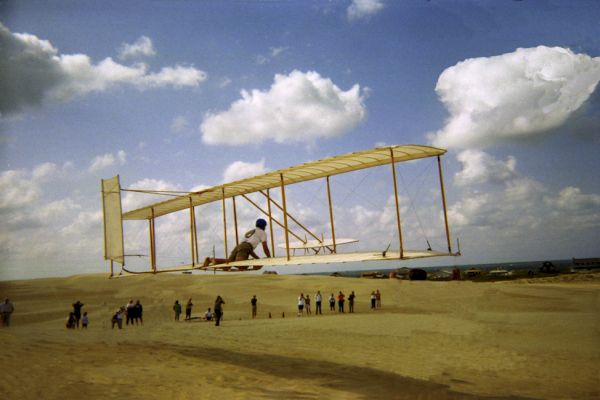 A person flies a replica of an early aircraft over a sandy expanse, with a group of people watching below, under a sky with scattered clouds.