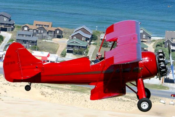 A red biplane is flying over a coastal area with sandy dunes and several houses, as well as a nearby ocean.