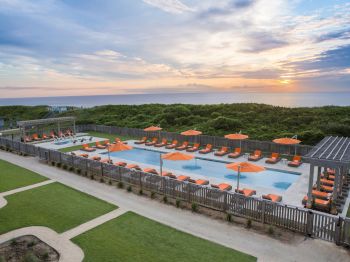 A serene outdoor pool area with orange umbrellas and lounge chairs overlooking a lush landscape and ocean under a partly cloudy sky at sunset.