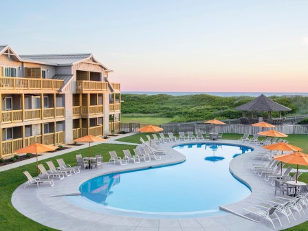 The image shows a resort with a kidney-shaped pool framed by lounge chairs and orange umbrellas, surrounded by grassy fields and a multi-story building.