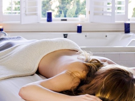 A woman lies face-down on a massage table covered with a towel in a spa-like bathroom setting with lit candles surrounding the bathtub.