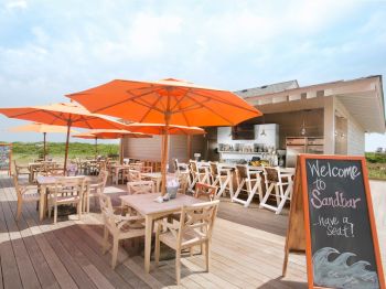 An outdoor seating area with wooden tables and chairs, shaded by orange umbrellas next to a bar. A chalkboard sign says, 