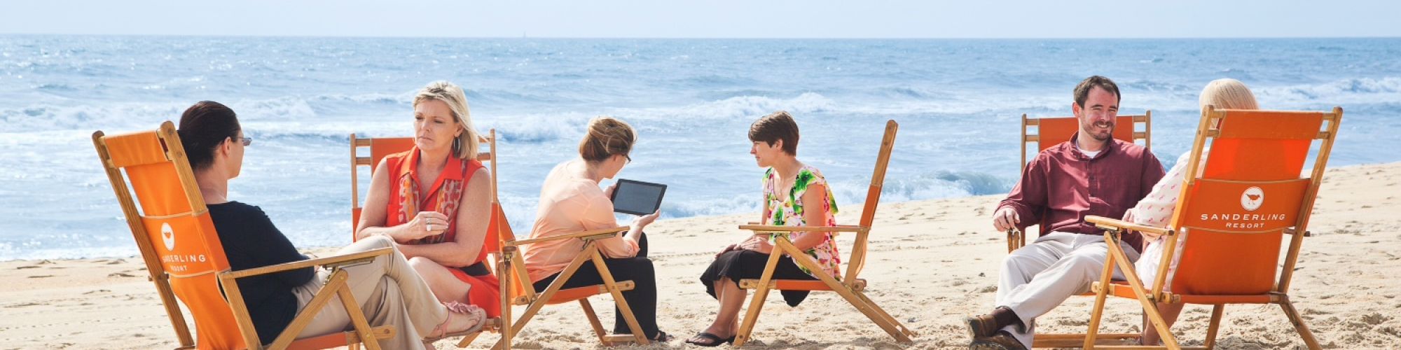 A group of people is sitting on orange beach chairs having conversations and using a laptop near the ocean on a sandy beach.