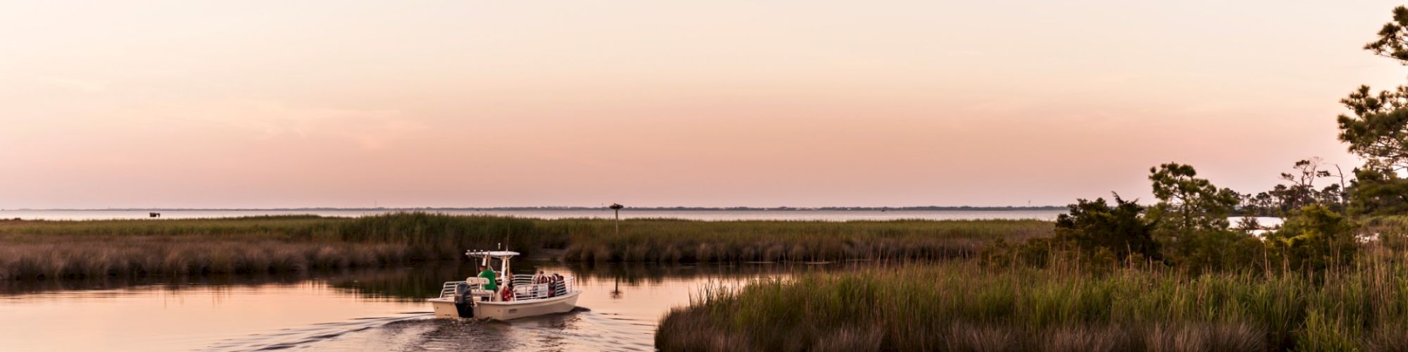 A small boat travels along a narrow waterway bordered by tall grass and trees, under a calm sky with a gentle sunset glow in the background.