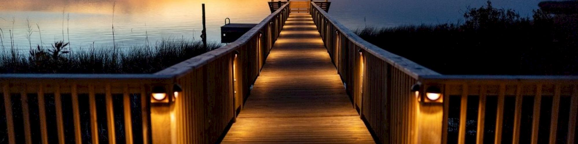 A wooden pathway, illuminated by lights, leads to a pavilion over tranquil water at sunset. The sky displays a beautiful gradient of colors.