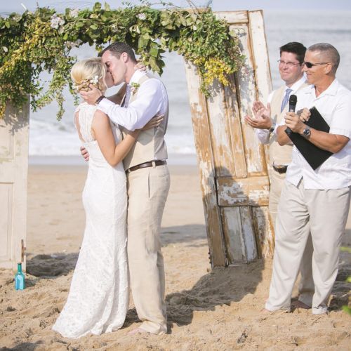 A couple is kissing at a beach wedding ceremony under an arch with greenery, with the officiant and wedding party members clapping nearby.