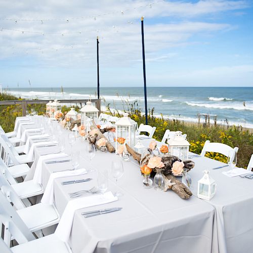 This image shows a long table set for a meal by the ocean. The table has white chairs, white tablecloth, and flower arrangements. The sentence ends.