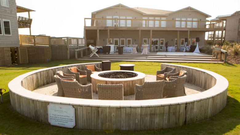 An outdoor seating area with wicker chairs surrounding a central fire pit, in front of a large, two-story house with a wraparound deck.