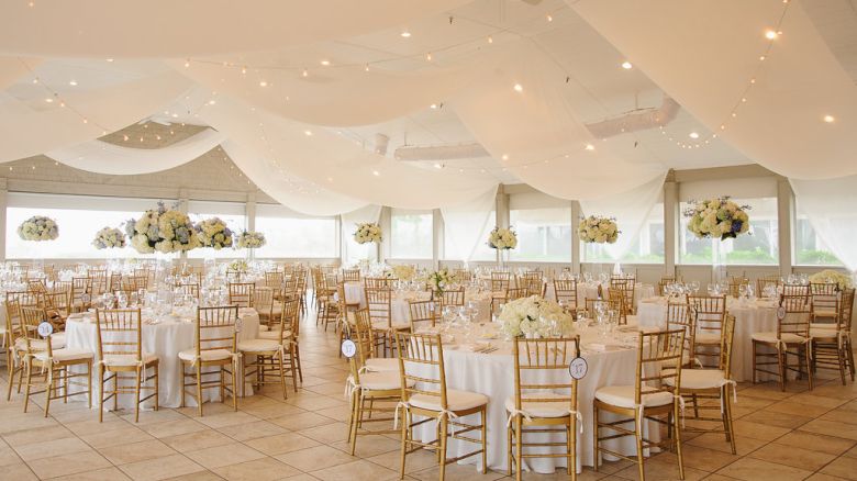 The image shows an elegantly decorated banquet hall with round tables, white tablecloths, gold chairs, and floral centerpieces, ready for a wedding or event.