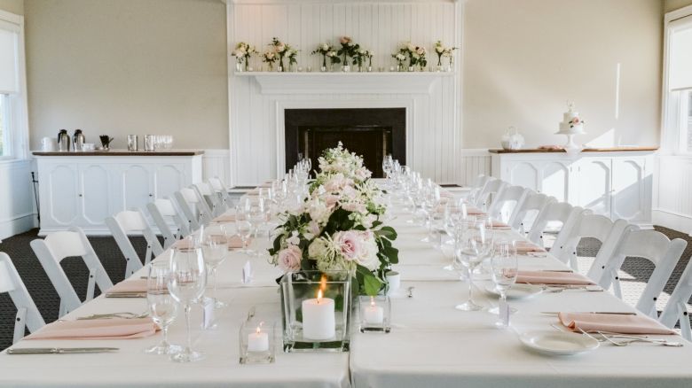 An elegantly set table with white linens, floral centerpieces, and candles in a spacious room with white chairs and a decorated mantel.